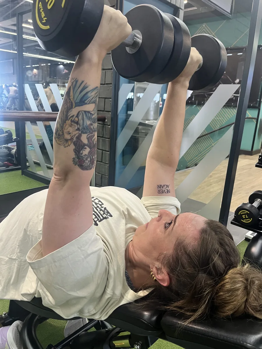 Megan pressing dumbbells, her “never again” tattoo visible